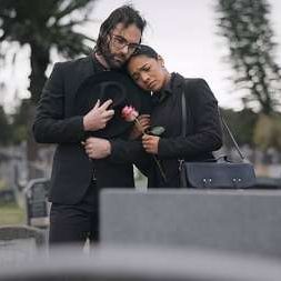Couple, sad and mourning at tomb of graveyard, funeral and pay respect together outdoor. Death, grief and man and woman at cemetery by casket, empathy and interracial support to console at tombstone.